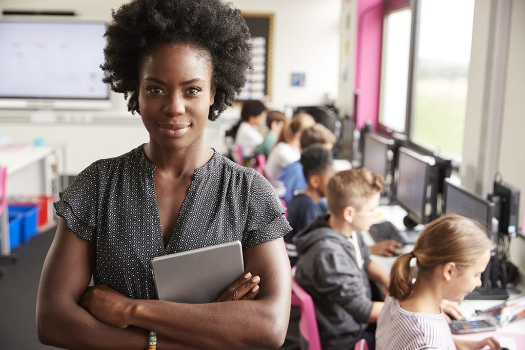 Retrato de uma professora segurando uma mesa digital e ensinando uma linha de alunos do ensino médio sentados em uma tela na sala de aula do computador.